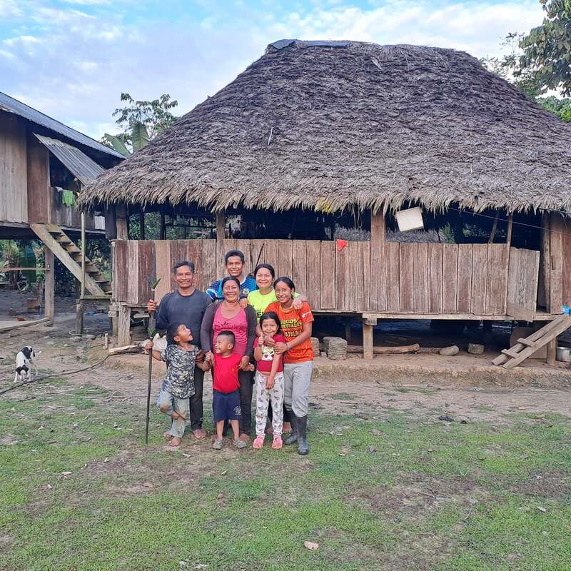 Un groupe de personnes souriantes, adultes et enfants, se tient devant une maison rustique en bois au toit de chaume, dans un village.
