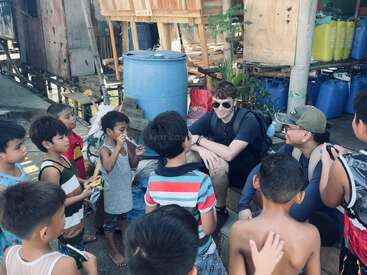 Un grupo de niños sonrientes se reúnen en torno a dos hombres sentados, uno de ellos con gafas de sol, en un entorno rural al aire libre con casas de madera, barriles y objetos dispersos.