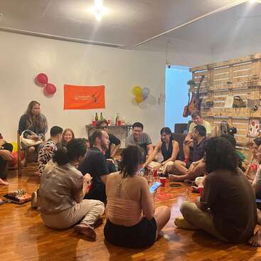 Un grupo de personas se reúne en el interior, sentadas en el suelo formando un círculo. Hablan, ríen y toman un aperitivo. La decoración incluye globos, bebidas y guitarras.