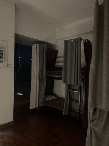 Esta imagen muestra una habitación de residencia universitaria con poca luz, con literas con cortinas, suelo de madera, una ventana que deja entrever la noche exterior y un aparato de aire acondicionado en la pared.