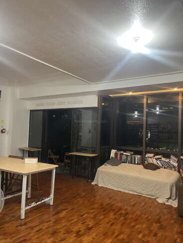 Esta imagen muestra una habitación acogedora y bien iluminada con suelo de madera, un sofá cubierto de libros, varias mesas y sillas, grandes ventanas y la frase «MÁS QUE SIMPLES LUGARES».