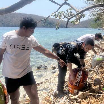 Three people are cleaning a rocky, littered beach under a sunny sky. They collect trash in bags beside clear blue water and scenic, tree-covered hills.