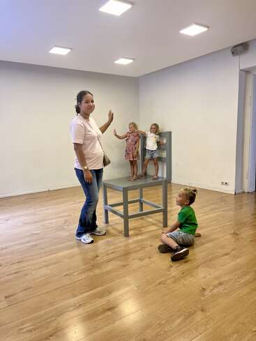 Une femme se tient à côté d'une grande chaise. Deux enfants apparaissent minuscules sur la chaise, et un autre enfant est assis sur le sol, créant ainsi une illusion d'optique.