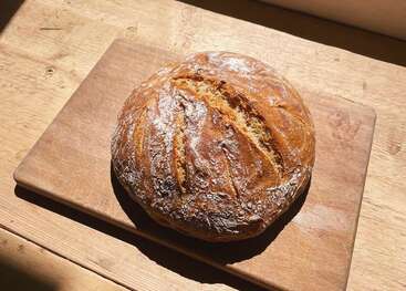 A round, rustic loaf of bread sits on a wooden cutting board in sunlight, its crust golden-brown with cracks, lightly dusted with flour, invitingly fresh.