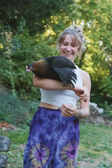 A young woman smiles joyfully outdoors, holding a black and brown chicken on her arm. She wears a white crop top and colorful tie-dye pants. Lush greenery surrounds them.