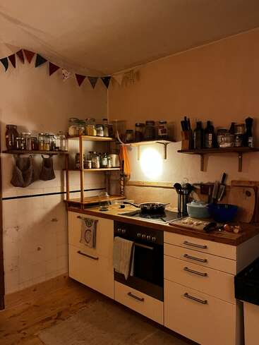 A cozy, dimly lit kitchen with wooden countertops, shelves of jars and spices, cooking utensils, colorful bunting, and a stove with food being prepared. Warm atmosphere.