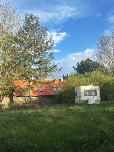 A white camper sits in tall green grass, backed by a rustic brick house with a red roof, surrounded by tall trees under a bright blue sky.