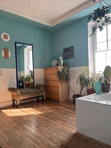 Sunlight fills a cozy, teal-walled room with wooden floors. There's a dresser, large mirror, plants, a patterned bench, wall art, and a white bathtub.