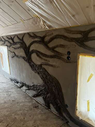 A textured tree mural is sculpted onto a wall in a room under construction, with plastic sheets protecting the ceiling and window frames. The branches spread widely.