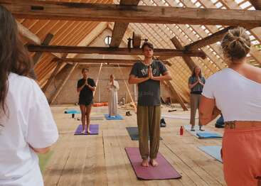 A group of people practice yoga on colorful mats in a rustic, wooden attic with exposed beams and natural light streaming through the roof. Calm atmosphere.