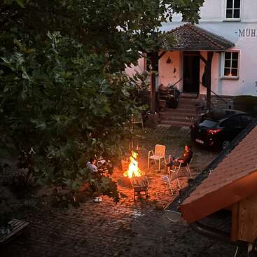 Two people sit around a bright fire on a cobblestone courtyard, surrounded by empty chairs and trees, in front of a house with steps and parked car.
