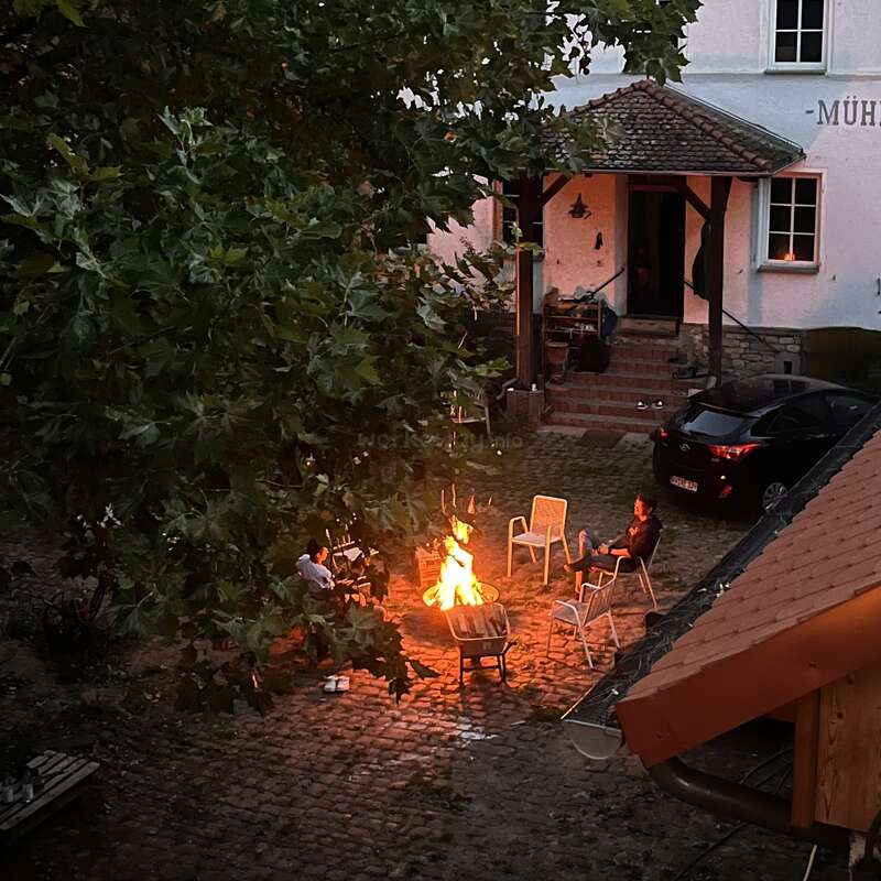 Two people sit around a bright fire on a cobblestone courtyard, surrounded by empty chairs and trees, in front of a house with steps and parked car.