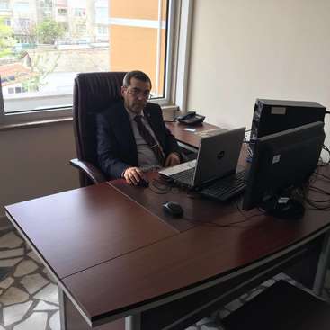 L'image représente un homme en costume assis à un bureau avec un ordinateur portable et un écran d'ordinateur, dans un bureau avec une fenêtre et une vue sur la ville.