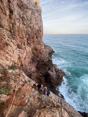 Zwei Menschen klettern an einem Seil auf eine zerklüftete Felsklippe am Meer, umgeben von blauen Wellen und einer dramatischen Landschaft. Ein atemberaubendes Abenteuer.