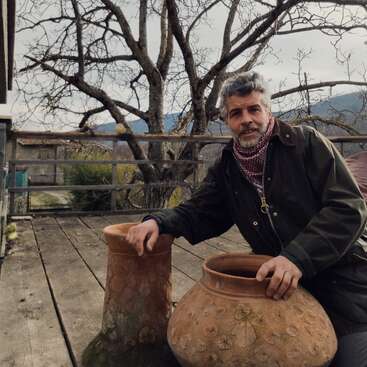 Un hombre con el pelo gris y una bufanda está sentado al aire libre en una terraza de madera, apoyado en dos grandes vasijas de barro decoradas. Detrás hay un árbol sin hojas.