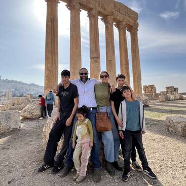 Una familia de seis miembros posa delante de unas antiguas columnas de piedra en un día soleado, con unas ruinas y un cielo azul al fondo. Todos parecen felices.