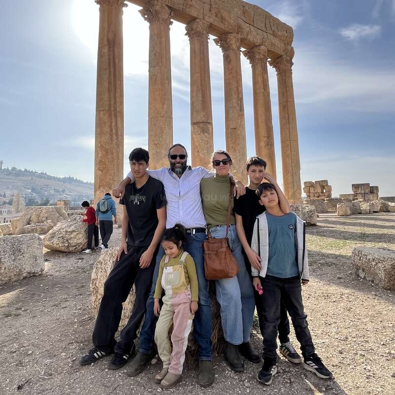 Une famille de six personnes pose devant d'anciennes colonnes de pierre par une journée ensoleillée, avec des ruines et un ciel bleu en arrière-plan. Tout le monde semble heureux.