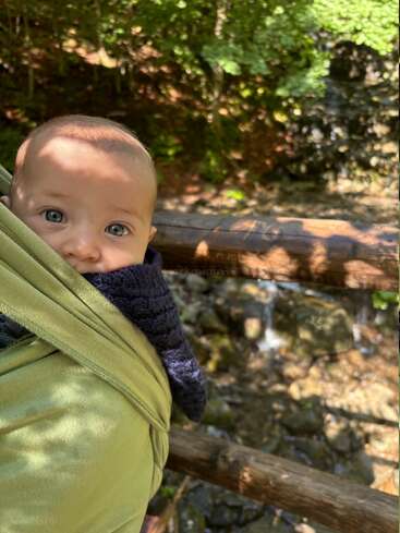 A baby with bright blue eyes is snuggled in a green wrap, outdoors by a wooden fence, with greenery and sunlight creating a peaceful atmosphere.