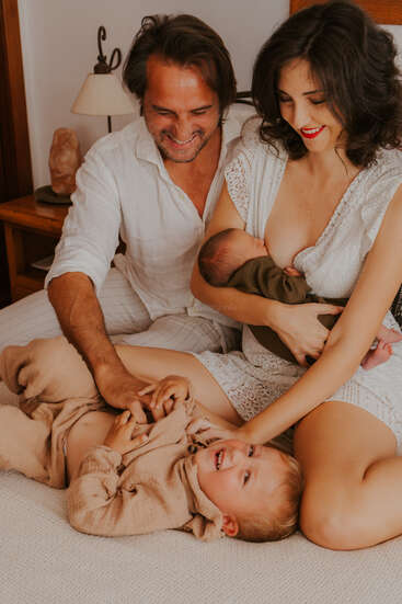 A joyful family shares a tender moment. The mother breastfeeds a newborn, the father plays with their laughing child, and everyone looks happy and content together.