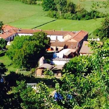 The image depicts a serene rural landscape, featuring a large white building with a brown roof, surrounded by lush greenery and trees, set against a picturesque countryside backdrop.