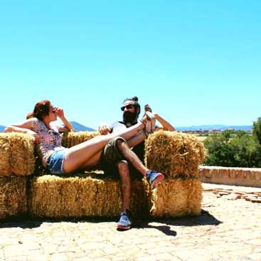 A man and woman sit on a couch made of hay bales, set against a backdrop of mountains and a clear blue sky on a sunny day.