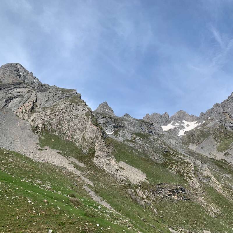 Dieses Bild zeigt schroffe Berggipfel mit Schneeflecken, grüne Grashänge, felsiges Gelände und einen strahlend blauen Himmel mit zarten Wolken, was eine friedliche Landschaft ergibt.
