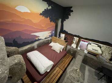 A cozy bedroom with a unique stone and wood design, mural of a scenic sunset landscape, soft bed, and an open view into a connected living space.