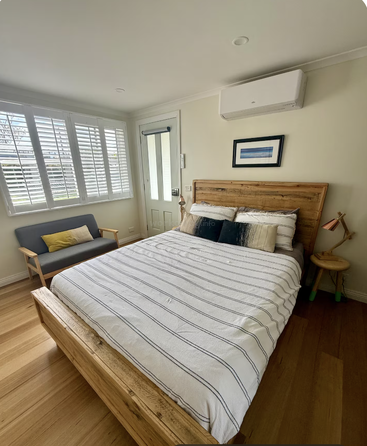 Dieses gemütliche Schlafzimmer hat ein Holzbett, gestreifte Bettwäsche, 'ne Bank mit bunten Kissen, Holzböden, Kunstwerke, 'nen Beistelltisch und große, helle Fenster.