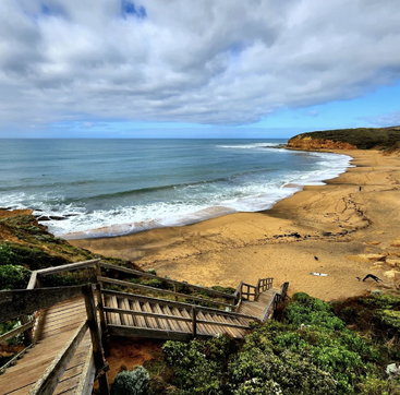 Eine Holztreppe führt runter zu einem riesigen Sandstrand mit sanften Wellen, dramatischen Klippen, üppigem Grün und einem wolkenverhangenen blauen Himmel über dem ruhigen Meer.