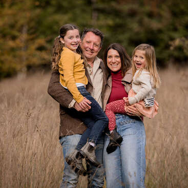 Una feliz familia de cuatro miembros posa junta en un campo cubierto de hierba. Los padres sostienen a sus hijas sonrientes. Visten ropa informal de otoño y parecen felices al aire libre.