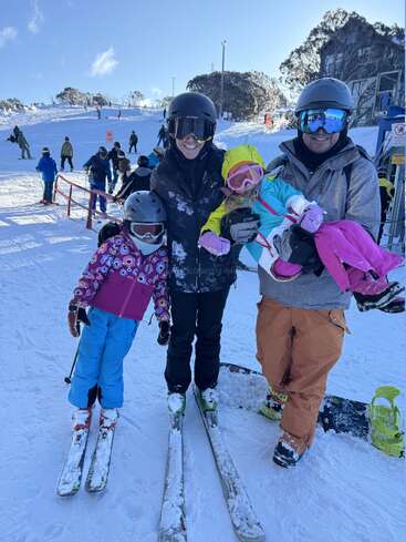 Eine Familie in bunten Skiklamotten posiert fröhlich auf einer verschneiten Piste. Die Eltern und ihre zwei Kinder sind bereit zum Skifahren und Snowboarden unter strahlend blauem Himmel.