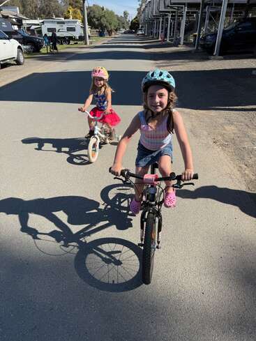 Duas meninas andam de bicicleta numa rua ensolarada, usando capacetes. As sombras delas se estendem pela calçada, cercadas por carros estacionados e prédios.
