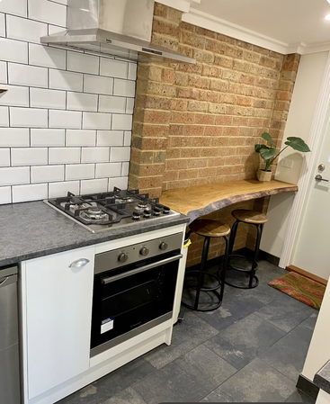 Essa cozinha tem um fogão moderno, forno, azulejos brancos tipo metrô, parede de tijolos à vista, balcão de madeira com borda viva, dois bancos e um vaso de planta.
