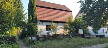 Une maison pittoresque au toit de tuiles rouges, entourée d'une végétation luxuriante et d'une clôture en bois. Une camionnette blanche garée et de l'herbe envahissante complètent cette scène paisible.