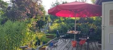 Un patio confortable en bois, entouré d'une végétation luxuriante, comprend des chaises éparpillées, une table avec des objets et un grand parasol rouge qui offre de l'ombre lors des journées ensoleillées.