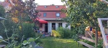 Une maison confortable au toit rouge se trouve au milieu d'une végétation luxuriante, d'arbres fruitiers, d'un jardin et d'un banc en bois, créant ainsi une oasis paisible et accueillante dans l'arrière-cour.