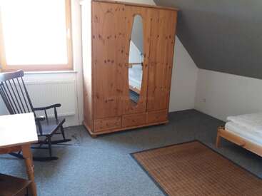 Ein einfaches, gemütliches Schlafzimmer im Dachgeschoss mit einem Kleiderschrank aus Holz und Spiegel, einem Einzelbett, einem Schaukelstuhl, einem Schreibtisch, einem Fenster, einem Teppich und einer schrägen Decke, das eine ruhige Atmosphäre schafft.