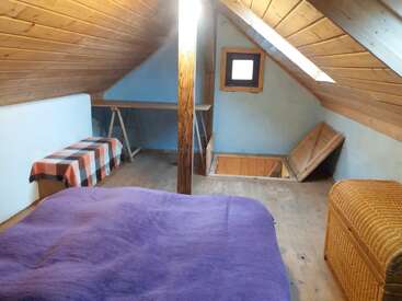 Dieses gemütliche Dachzimmer hat Holzdecken, ein lila Bett, eine karierte Bank, eine Weidenkommode, einen kleinen Schreibtisch und eine Luke für die Treppe.