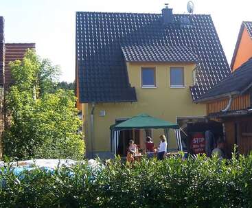 Ein gelbes Haus mit einem dunkelgrauen Dach steht hinter einer Hecke. Vier Leute hängen unter einem grünen Sonnendach im sonnigen Garten ab und quatschen miteinander.