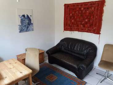 In diesem Zimmer gibt's ein schwarzes Ledersofa, einen blauen Teppich, zwei beige Stühle, einen Holztisch, ein Zebra-Gemälde und einen roten Wandbehang.
