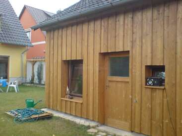 Auf dem Bild sieht man einen kleinen Holzschuppen mit einem Fenster, einer Tür und einem Regal, auf dem ein Radio steht. Auf dem Rasen liegen ein Gartenschlauch und eine Gießkanne.
