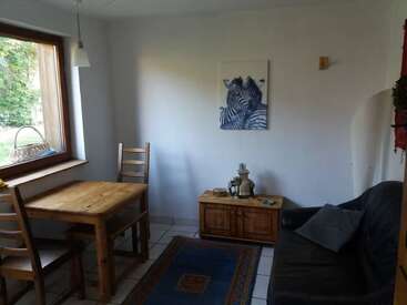 Ein gemütliches Zimmer mit einem Holztisch, zwei Stühlen, einem schwarzen Sofa, einem Schrank, Zebra-Wandkunst, einem Fenster, einem blauen Teppich und sanftem Tageslicht.