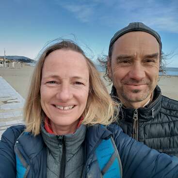 Una mujer y un hombre sonrientes se hacen un selfie juntos en una playa, con chaquetas. El cielo es azul, hace frío y la playa está vacía.