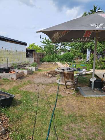 Um jardim nos fundos com canteiros elevados, um grande guarda-sol protegendo uma mesa e cadeiras de madeira, ferramentas de jardinagem espalhadas, uma mangueira e alguns materiais de construção, sob um céu parcialmente nublado.