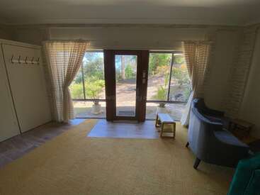 Esta acogedora sala de estar cuenta con una puerta de cristal, grandes ventanales con cortinas ligeras, alfombra amarilla, sillón, percheros, mesitas y exuberante vegetación exterior a la vista.