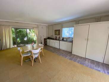 Esta imagen muestra una sala diáfana y luminosa con mesa de comedor y cuatro sillas, cocina americana, grandes ventanales, puerta corredera de cristal y suelo de moqueta beige.