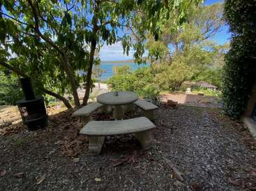 Bajo frondosos árboles hay una mesa de picnic de piedra con bancos curvados. Cerca hay una rústica chimenea al aire libre, con vistas a una exuberante vegetación y a un sereno lago azul.