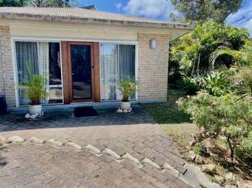 Una casita de ladrillo con grandes puertas y ventanas de cristal, flanqueada por dos macetas, rodeada de vegetación y un camino pavimentado bajo un cielo soleado.
