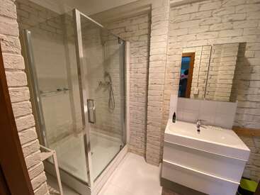 Este cuarto de baño cuenta con una cabina de ducha de cristal, un moderno tocador blanco con cajones, un armario con espejo, paredes de ladrillo blanco visto, suelo de baldosas y una iluminación clara y limpia en toda la estancia.