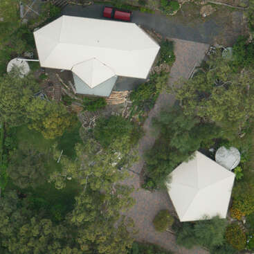 The image depicts an aerial view of two white houses with octagonal roofs, surrounded by trees and a paved driveway, situated in a rural area.
